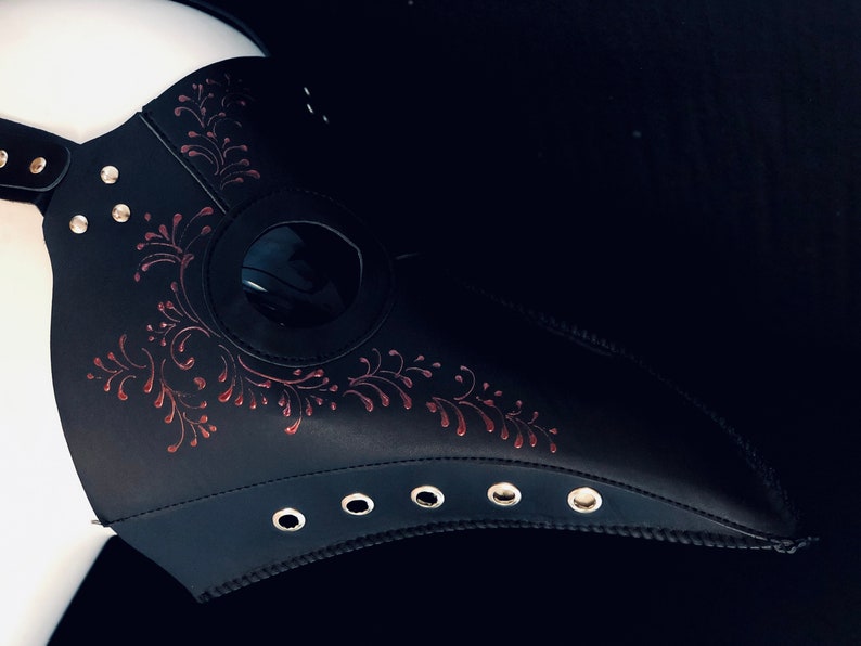 Puede incluir: Una m&aacute;scara de m&eacute;dico de la peste de cuero negro con un dise&ntilde;o floral rojo y una pieza ocular transparente. La m&aacute;scara tiene un pico largo y se asegura con correas.