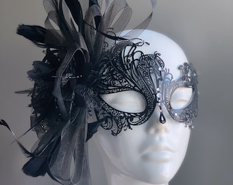 May include: A black masquerade mask with intricate lace detailing and rhinestones. The mask is attached to a black feather and lace headpiece.