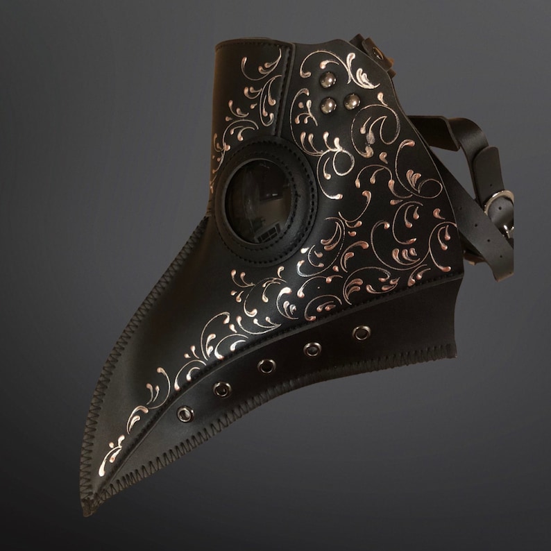 May include: A black leather plague doctor mask with silver floral details and a clear plastic eye piece. The mask has a strap that goes around the back of the head.