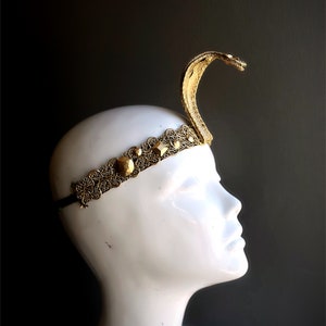 May include: A gold-colored cobra headpiece with an ornate filigree headband. The snake's head is raised, and the headband features geometric accents. The headpiece is displayed on a white mannequin head against a dark background.