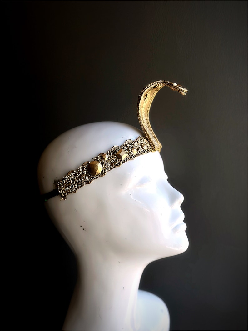 May include: A gold-colored cobra headpiece with an ornate filigree headband. The snake's head is raised, and the headband features geometric accents. The headpiece is displayed on a white mannequin head against a dark background.