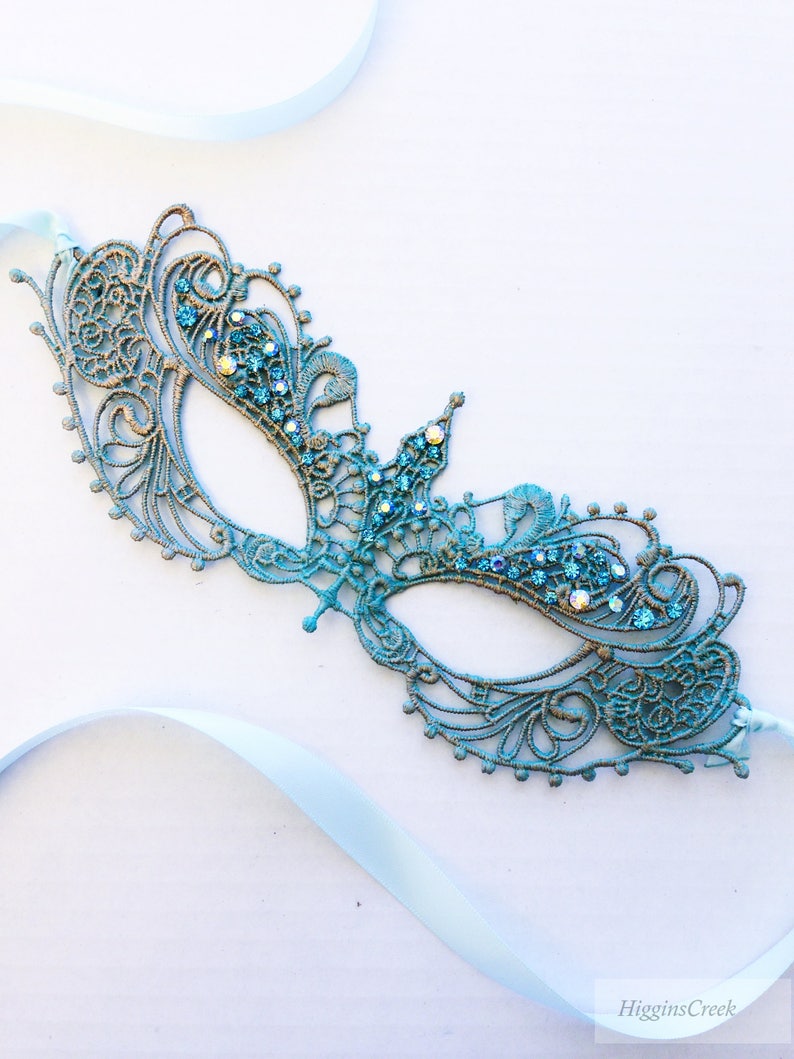 May include: A teal blue masquerade mask with intricate lace detailing and sparkling rhinestones. The mask is attached to a light blue satin ribbon.