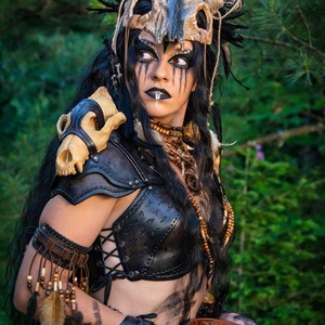 May include: A woman wearing a leather costume with a skull headdress and a skull shoulder piece. She is holding a wooden bowl filled with pine cones.