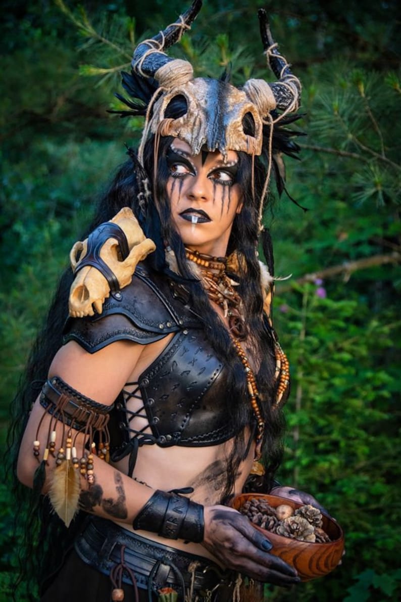 May include: A woman wearing a leather costume with a skull headdress and a skull shoulder piece. She is holding a wooden bowl filled with pine cones.