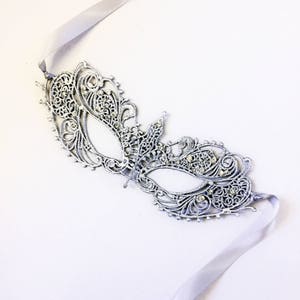 May include: A silver masquerade mask with intricate lace detailing and rhinestones. The mask is attached to a silver ribbon.
