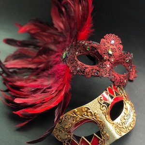 Puede incluir: Dos máscaras decorativas de carnaval. La máscara superior es roja con detalles de encaje, gemas y plumas rojas. La máscara inferior es dorada, roja y negra con un patrón de arlequín y notas musicales.