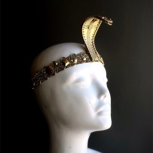 May include: A gold-colored headpiece featuring a cobra design. The cobra rises from a decorative band adorned with gemstones and intricate patterns. The headpiece is displayed on a white mannequin head against a dark background.