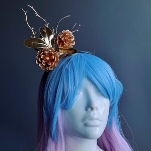 May include: A gold and brown hair accessory with pine cones and branches. The accessory is worn on a mannequin with blue and pink hair.