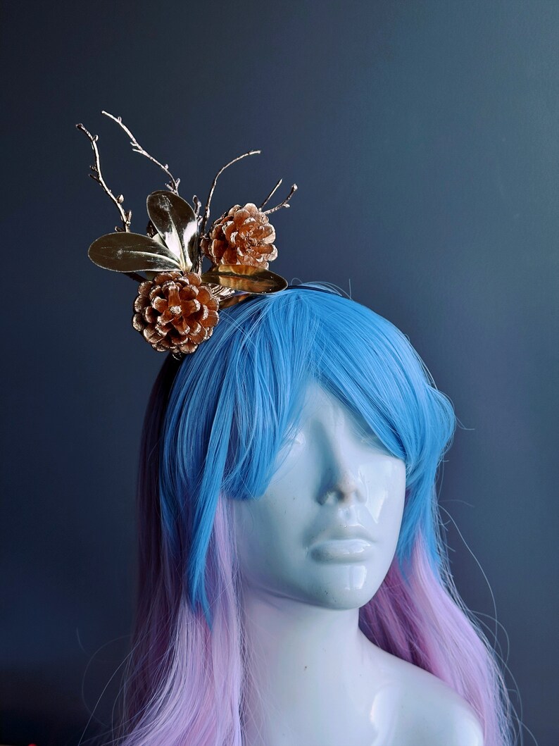 May include: A gold and brown hair accessory with pine cones and branches. The accessory is worn on a mannequin with blue and pink hair.