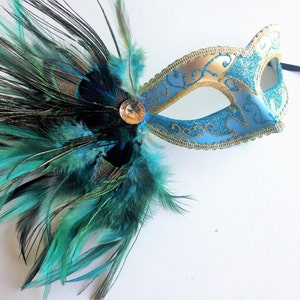 May include: A blue and gold masquerade mask with a peacock feather trim. The mask has a glitter finish and gold accents. The feather trim is a mix of teal and black feathers.