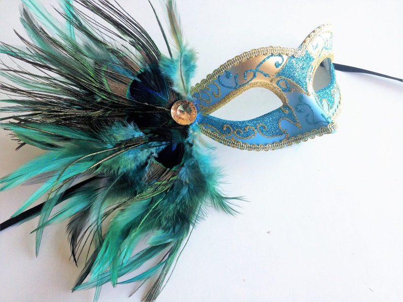 May include: A blue and gold masquerade mask with a peacock feather trim. The mask has a glitter finish and gold accents. The feather trim is a mix of teal and black feathers.
