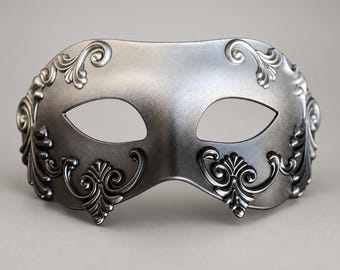 Può includere: Una maschera da ballo in maschera color argento con una finitura metallica liscia e dettagli intricati a volute in rilievo attorno alle aperture per gli occhi e ai bordi. La maschera ha una forma curva e simmetrica con due fori.