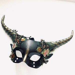 May include: A black and gold masquerade mask with large, spiraled horns. The mask is decorated with faux foliage and leaves in shades of green, pink, and gold. The horns are a bronze color.