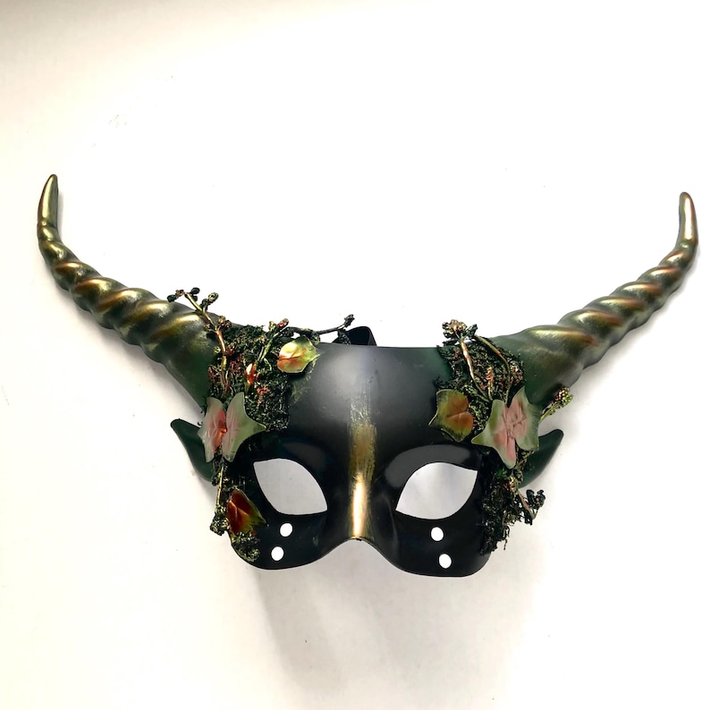 May include: A black masquerade mask with gold accents and two large, curved horns. The horns are a dark green with gold highlights. The mask is decorated with faux foliage and flowers. The eye holes are oval-shaped.