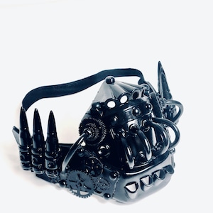 May include: A black steampunk mask with intricate detailing. The mask incorporates gear-like elements, bullet-shaped spikes, and a black elastic strap. The mask is crafted from a glossy, dark material, presenting a futuristic, industrial style.
