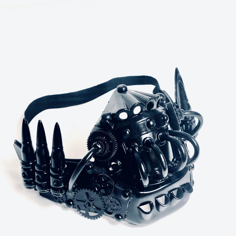 May include: A black steampunk mask with intricate detailing. The mask incorporates gear-like elements, bullet-shaped spikes, and a black elastic strap. The mask is crafted from a glossy, dark material, presenting a futuristic, industrial style.