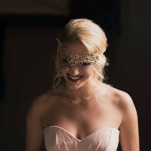 May include: A woman wearing a gold lace masquerade mask and a pink strapless dress. She is smiling and looking down.