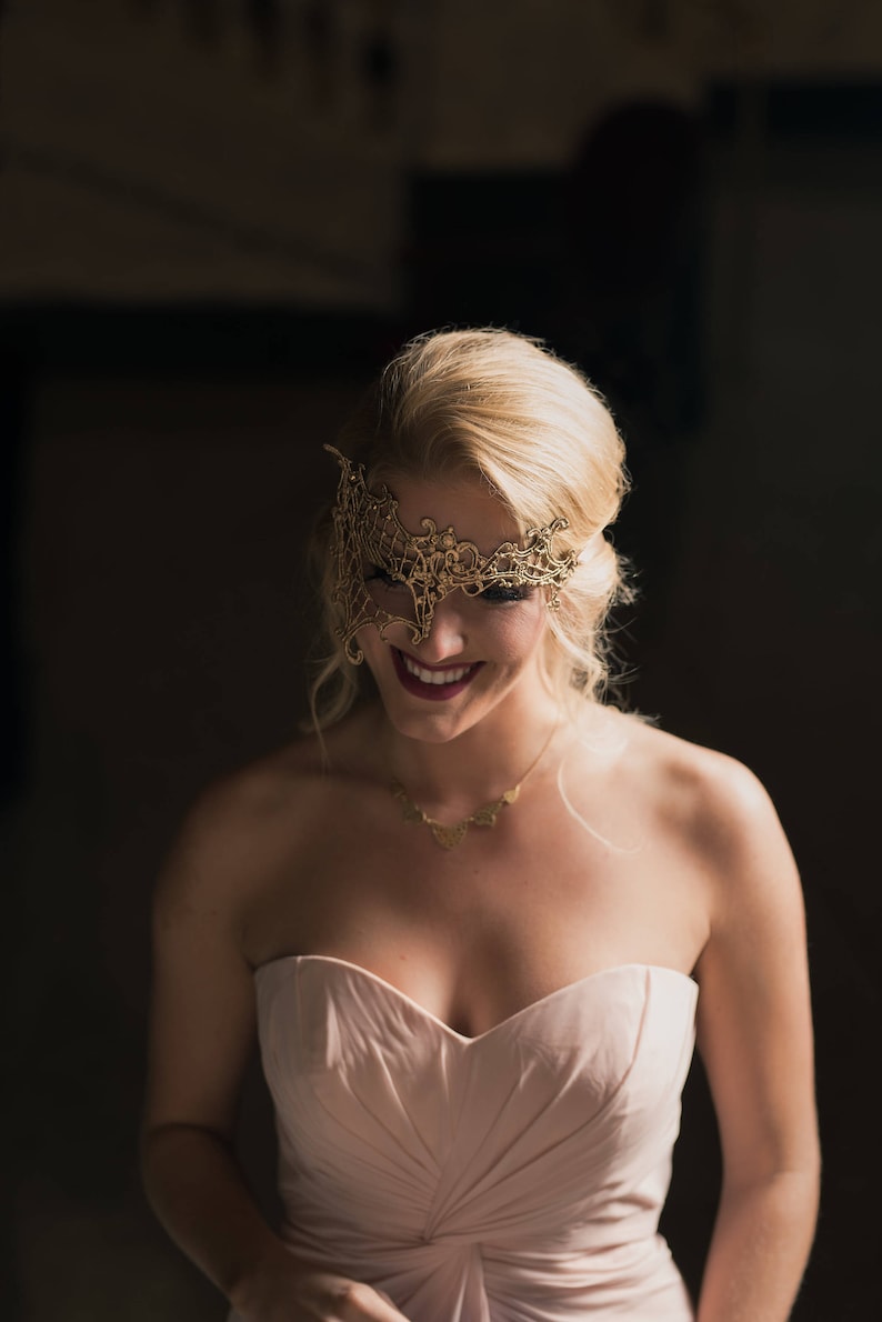 May include: A woman wearing a gold lace masquerade mask and a pink strapless dress. She is smiling and looking down.