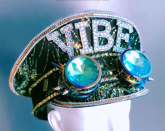 May include: Black and green sequined hat with the word "VIBE" spelled out in sparkling rhinestones. The hat features large, teal-colored goggles with silver spikes and a gold trim. The hat is displayed on a mannequin head.