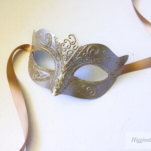 May include: A silver masquerade mask with gold glitter and intricate designs. The mask is attached to a long, thin, gold ribbon.