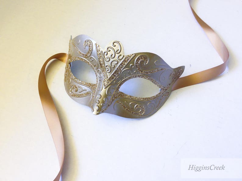 May include: A silver masquerade mask with gold glitter and intricate designs. The mask is attached to a long, thin, gold ribbon.