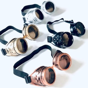 May include: A selection of steampunk goggles in silver, gold, bronze, and black. Each pair has round lenses and adjustable black elastic straps. The goggles have decorative details and a vintage aesthetic, reminiscent of Victorian era designs.
