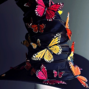 May include: A black witch's hat with a wide brim and a tall, pointed crown. The hat is decorated with a variety of colorful butterflies, including red, orange, yellow, and black.
