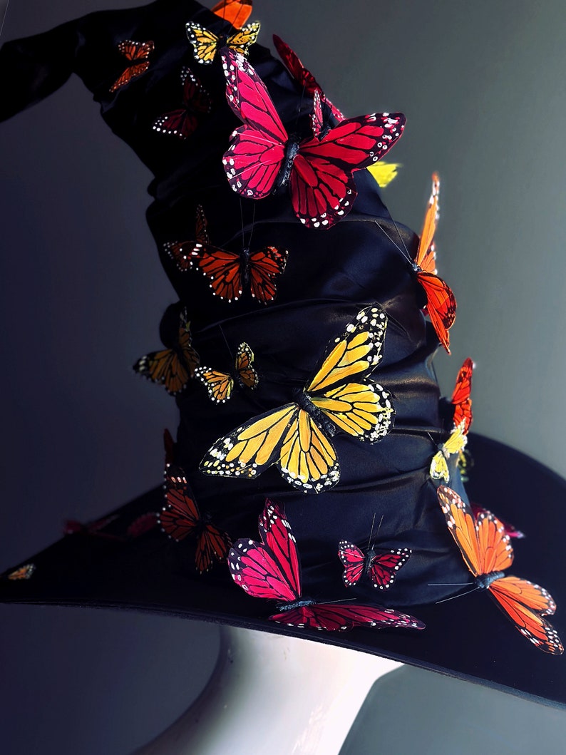 May include: A black witch's hat with a wide brim and a tall, pointed crown. The hat is decorated with a variety of colorful butterflies, including red, orange, yellow, and black.