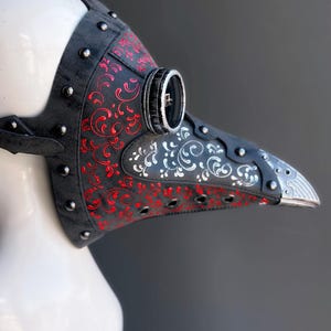 May include: A black plague doctor mask with red and white floral designs. The mask has a long, beak-like nose, silver accents, and is studded with silver rivets. The mask is made of leather and has a strap for securing it to the head.