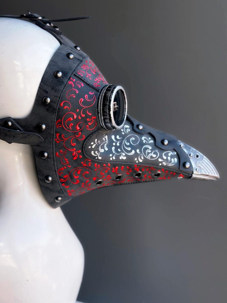 May include: A black plague doctor mask with red and white floral designs. The mask has a long, beak-like nose, silver accents, and is studded with silver rivets. The mask is made of leather and has a strap for securing it to the head.