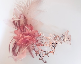 May include: A rose gold masquerade mask with a delicate, intricate design. The mask is adorned with rhinestones and features a feathered accent in shades of pink and brown.