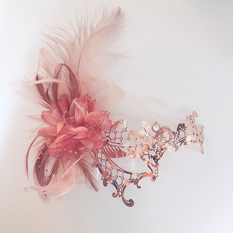 May include: A rose gold masquerade mask with a delicate, intricate design. The mask is adorned with rhinestones and features a blush-colored feather and flower accent.