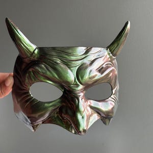 May include: A metallic green devil mask with two upward-curving horns. The mask has eye openings and a detailed, textured surface. The color shifts between green and bronze, creating a unique, iridescent effect. The mask is likely for costume or decorative purposes.