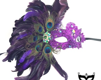 Puede incluir: Máscara de carnaval morada con un diseño de pluma de pavo real. La máscara está decorada con diamantes de imitación y una joya verde. Las plumas son negras y moradas.
