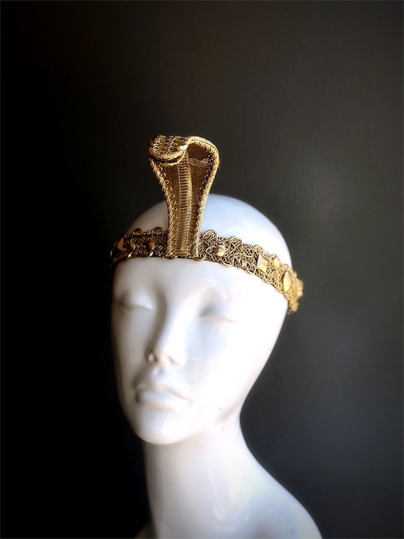 May include: A gold colored headband with a snake design. The headband has a decorative pattern and small round accents.
