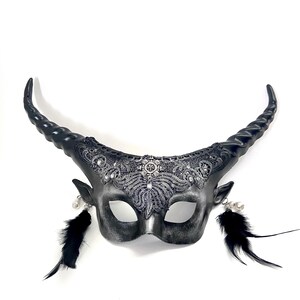 May include: A silver and black horned mask with intricate lace detailing and dangling black feathers. The mask features curved horns, small ear details, and a decorative center piece.
