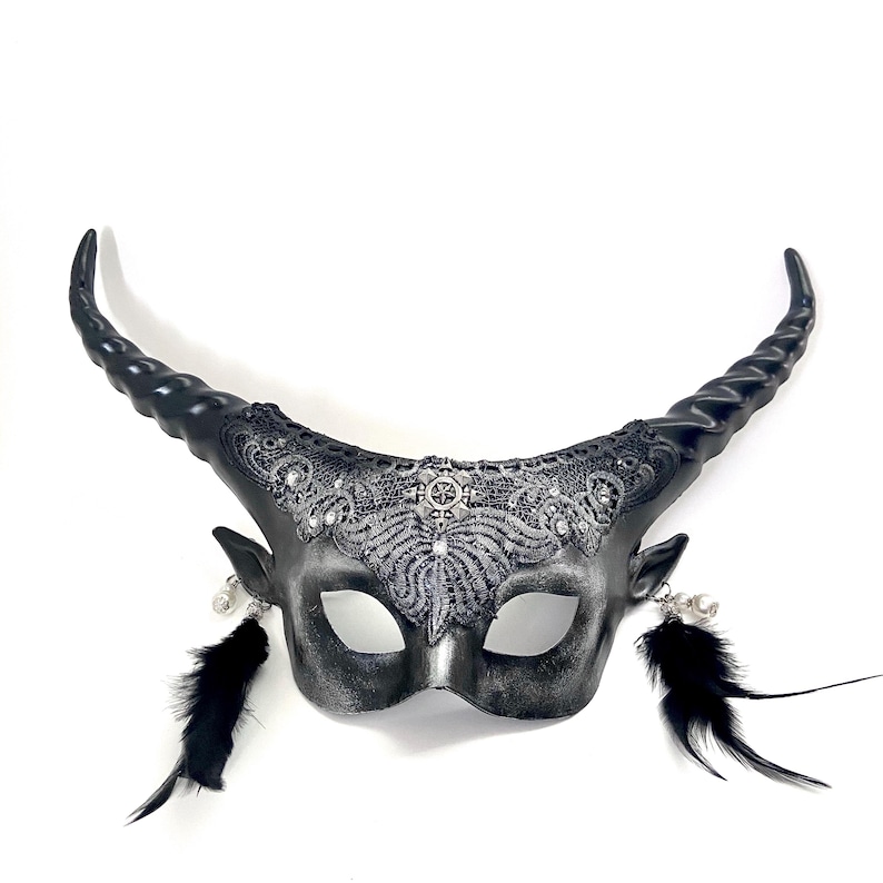 May include: A silver and black horned mask with intricate lace detailing and dangling black feathers. The mask features curved horns, small ear details, and a decorative center piece.