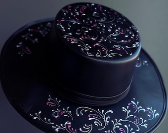 Puede incluir: Un sombrero de cuero negro con ala ancha y copa alta. El sombrero está decorado con un diseño floral blanco y rosa.