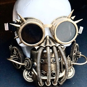 May include: A white skull adorned with steampunk-style goggles and a mask. The goggles are gold-toned with dark lenses and spiked details. The mask incorporates tubes and gears, all finished in a metallic bronze, offering a distinctive, industrial appearance.