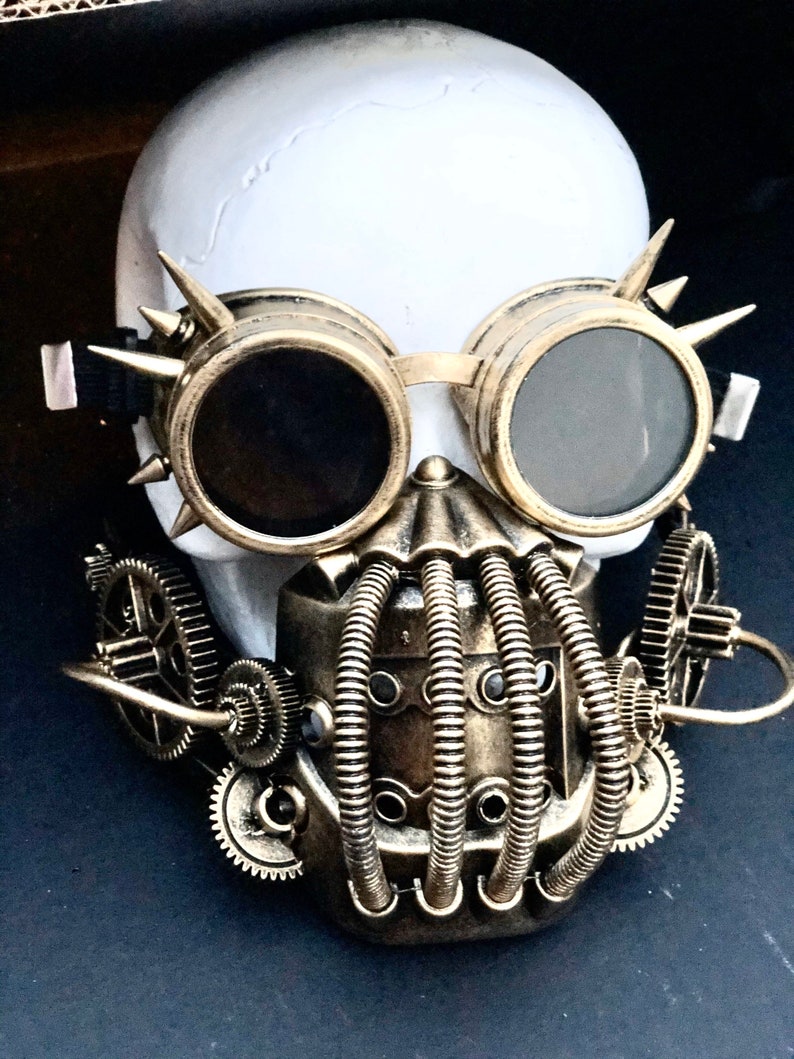 May include: A white skull adorned with steampunk-style goggles and a mask. The goggles are gold-toned with dark lenses and spiked details. The mask incorporates tubes and gears, all finished in a metallic bronze, offering a distinctive, industrial appearance.