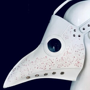 May include: White leather plague doctor mask with a long beak and a clear plastic eye piece. The mask has a pink floral design and metal eyelets around the edges.