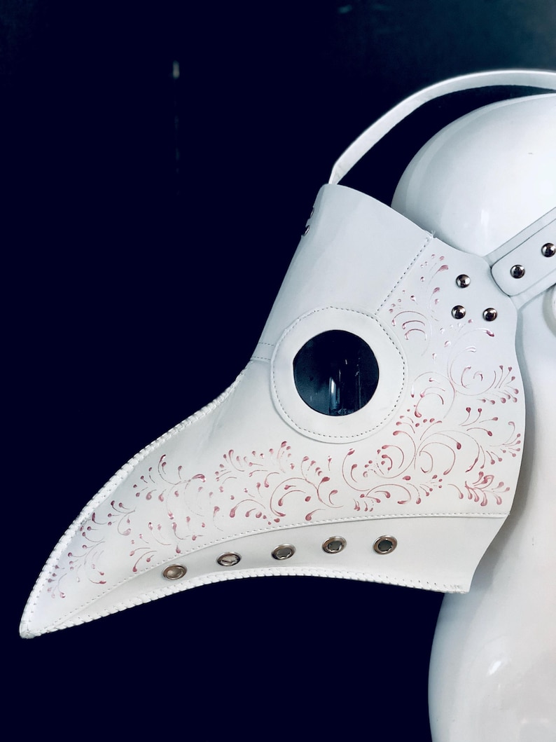 May include: White leather plague doctor mask with a long beak and a clear plastic eye piece. The mask has a pink floral design and metal eyelets around the edges.