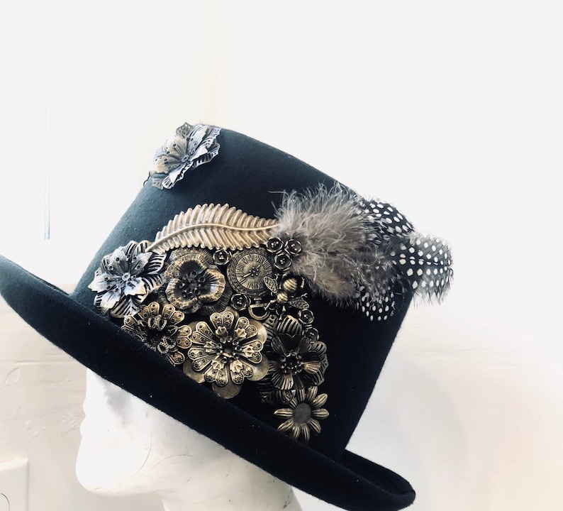 May include: A black top hat with a wide brim and a tall crown, decorated with gold and silver floral embellishments and a black and white feather.
