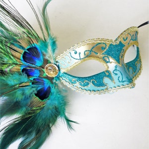 May include: A teal and gold masquerade mask with a peacock feather accent. The mask has a delicate gold trim and intricate designs.