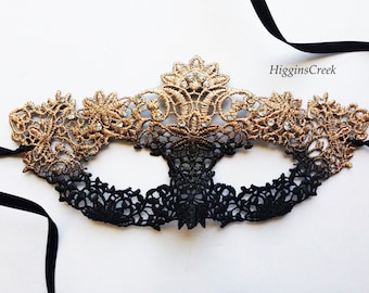 Può includere: Una maschera da ballo in pizzo nero e oro con strass. La maschera ha una parte inferiore in pizzo nero e una parte superiore in pizzo oro con un effetto sfumato.