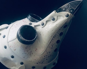 May include: A white plague doctor mask featuring a long, pointed beak and two circular eye lenses with dark, distressed silver rims. The surface is decorated with an intricate, swirling filigree pattern and secured with metallic rivets.