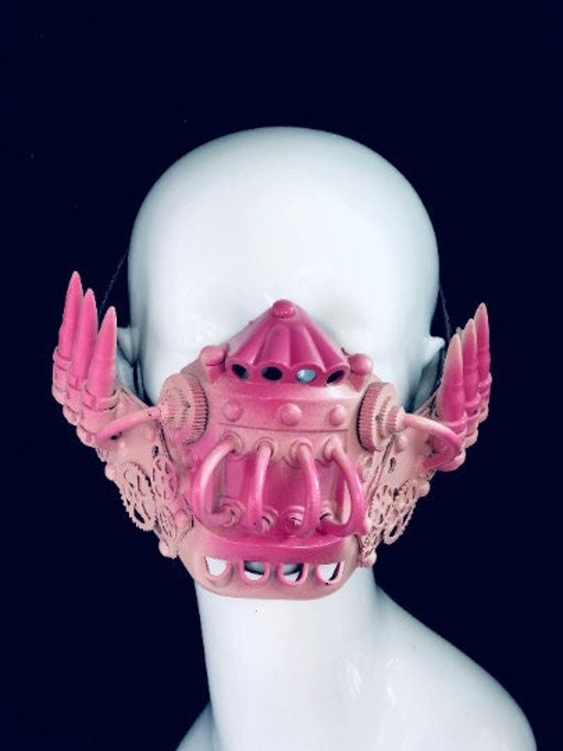 May include: A pink and beige steampunk-style mask with intricate details. The mask features a central, rounded structure with a series of curved tubes and gear-like elements. The mask is set against a dark background.