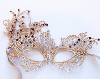 May include: A gold lace masquerade mask with purple rhinestones. The mask has a delicate, intricate design and is perfect for a masquerade ball or costume party.