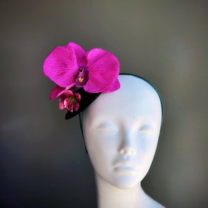 May include: A decorative headpiece featuring a vibrant fuchsia orchid and smaller pink flowers. The flowers are attached to a small black base with a green headband. The headpiece is displayed on a white mannequin head.