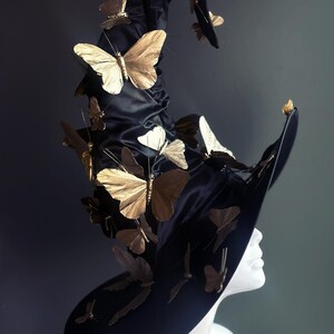 May include: A black witch hat adorned with numerous gold-colored butterfly embellishments. The hat has a wide brim and a tall, pointed crown. The butterflies are of varying sizes and are attached to the hat in a seemingly random pattern.